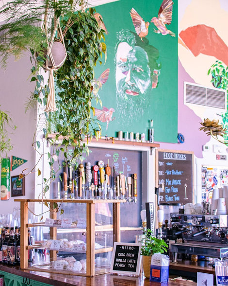 Photo of the interior of Grateful Brew. There is a pastry display case, a wall of beer taps, an espresso machine, plants on the bar and hanging from the ceiling, and the walls are painted with murals.