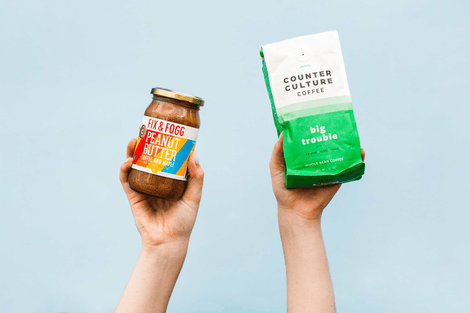 Photo of two hands in the air. One is holding a jar of Counter Culture Coffee x Fix & Fogg Maple Peanut Butter and the other is holding a bag of Big Trouble coffee.