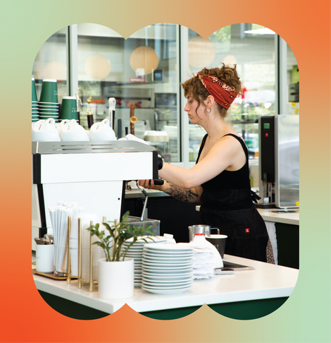 A barista in a red bandana and black tank top and apron prepares a drink on a white espresso machine.