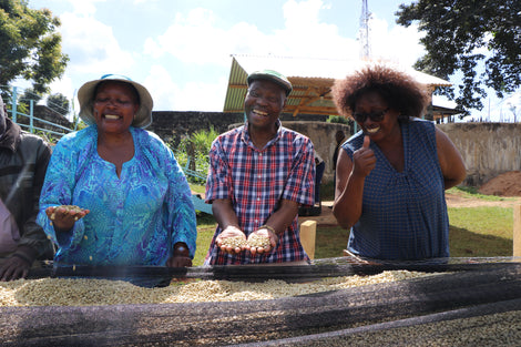 Photo of three members of Kushikamana holding coffee beans that are on raised drying racks.