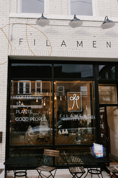 Photo of the exterior of Filament Coffee. There are signs on the windows that say, "Coffee, Tea, Plants, Good People" and "Filament Coffee & Tea"