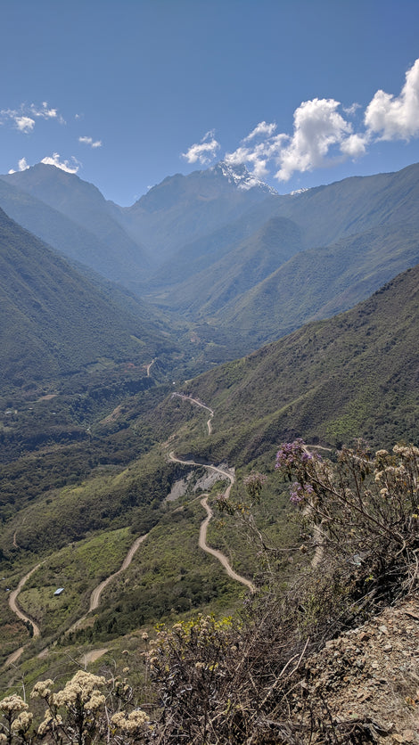 Photo looking down into the valley in Cusco, Peru. There are mountains covered in trees, winding dirt roads, and snow on a mountain peak in the distance.